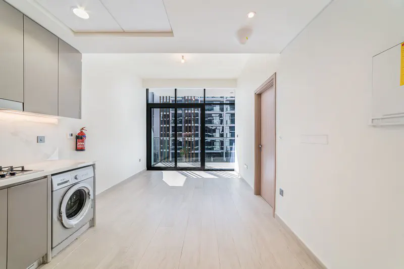 Modern kitchen area with light wood floors, grey cabinets, a built-in washing machine, and a balcony with glass doors.