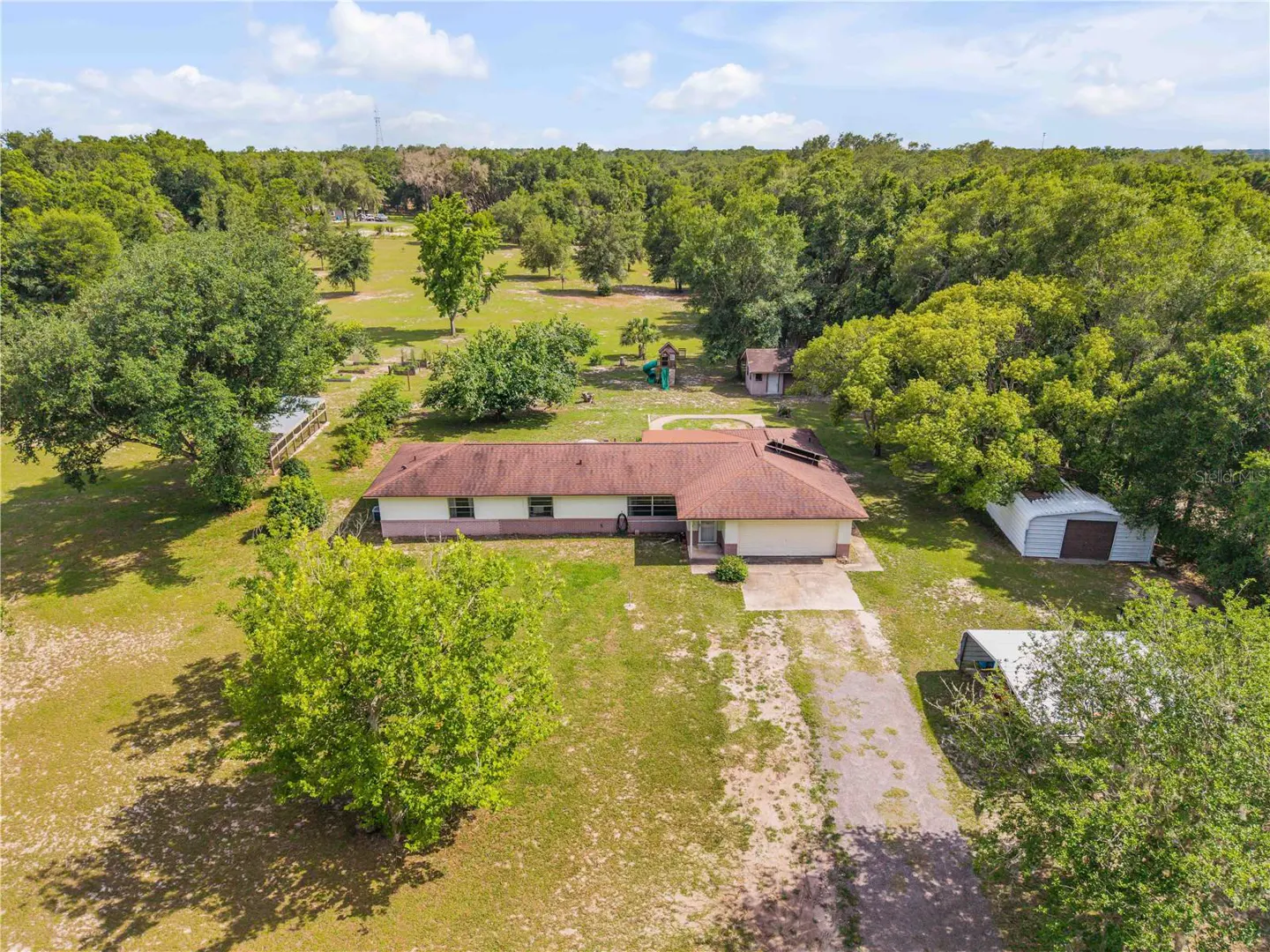Aerial view of a single-story home with a brown roof, large grassy yard, detached sheds, and surrounding forest.
