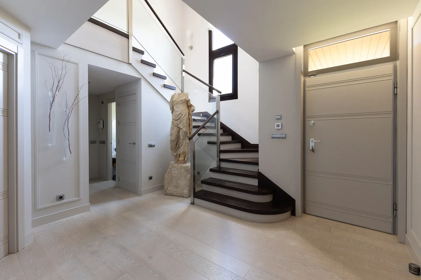 Modern foyer with light wood floors, a staircase with dark treads, glass railings, and a classical statue on a stone base.