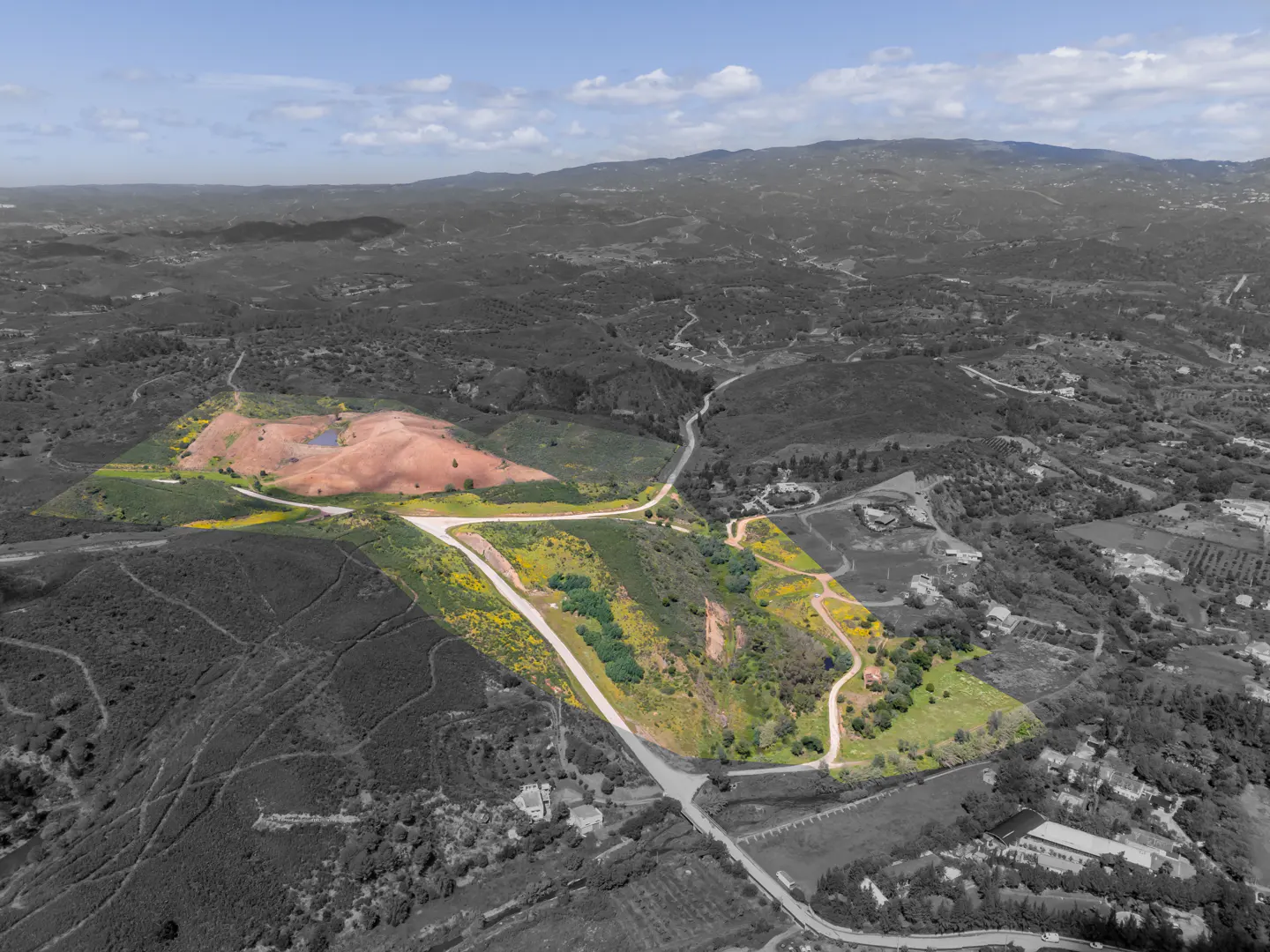 Aerial view of land with green and yellow vegetation, a red dirt patch with a pond, and a gray-scale background of hills and roads.
