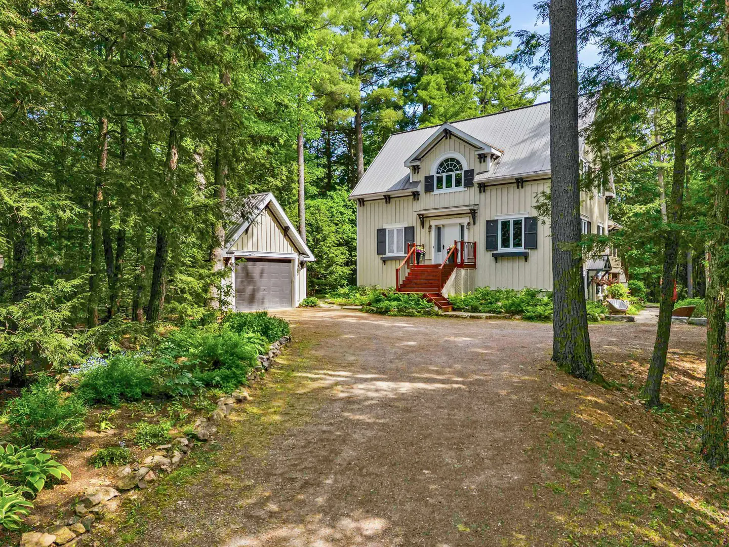 A light-colored two-story house with a metal roof and red stairs, set in a lush, wooded area with a gravel driveway.