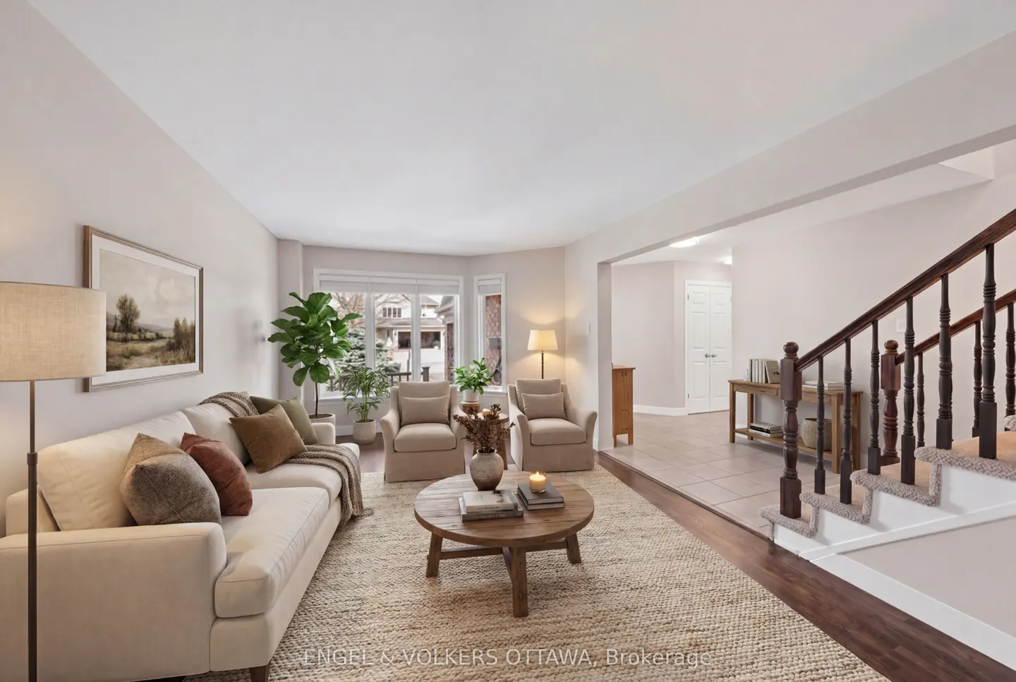 Living room with a beige sofa, two armchairs, and a round wooden coffee table on a woven rug. Staircase on the right.