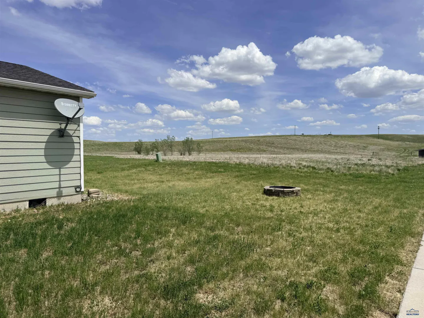 Backyard with a stone fire pit, green grass, and a view of open rolling hills under a blue sky with white clouds.