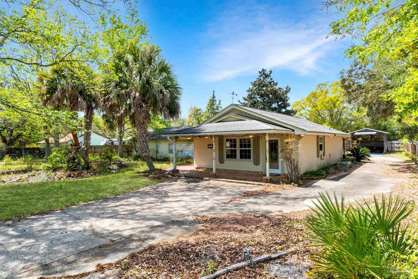 A tan single-story house with a covered porch and a long driveway on a sunny day. Palm trees and other greenery surround the house.