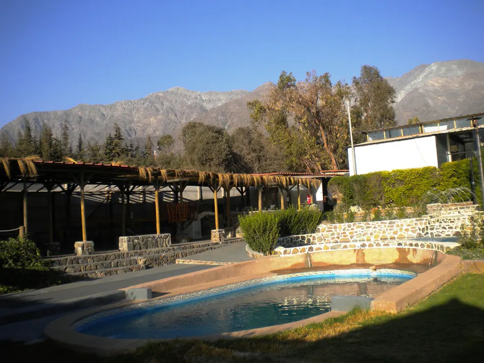 Outdoor pool with mountain backdrop. Covered patio with thatched roof. Stone accents and green landscaping. Blue sky.