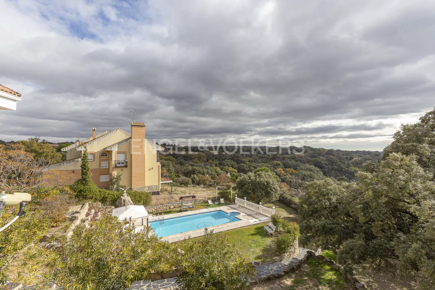 A high-angle view of a yellow house with a pool and a forest in the background under a cloudy sky.