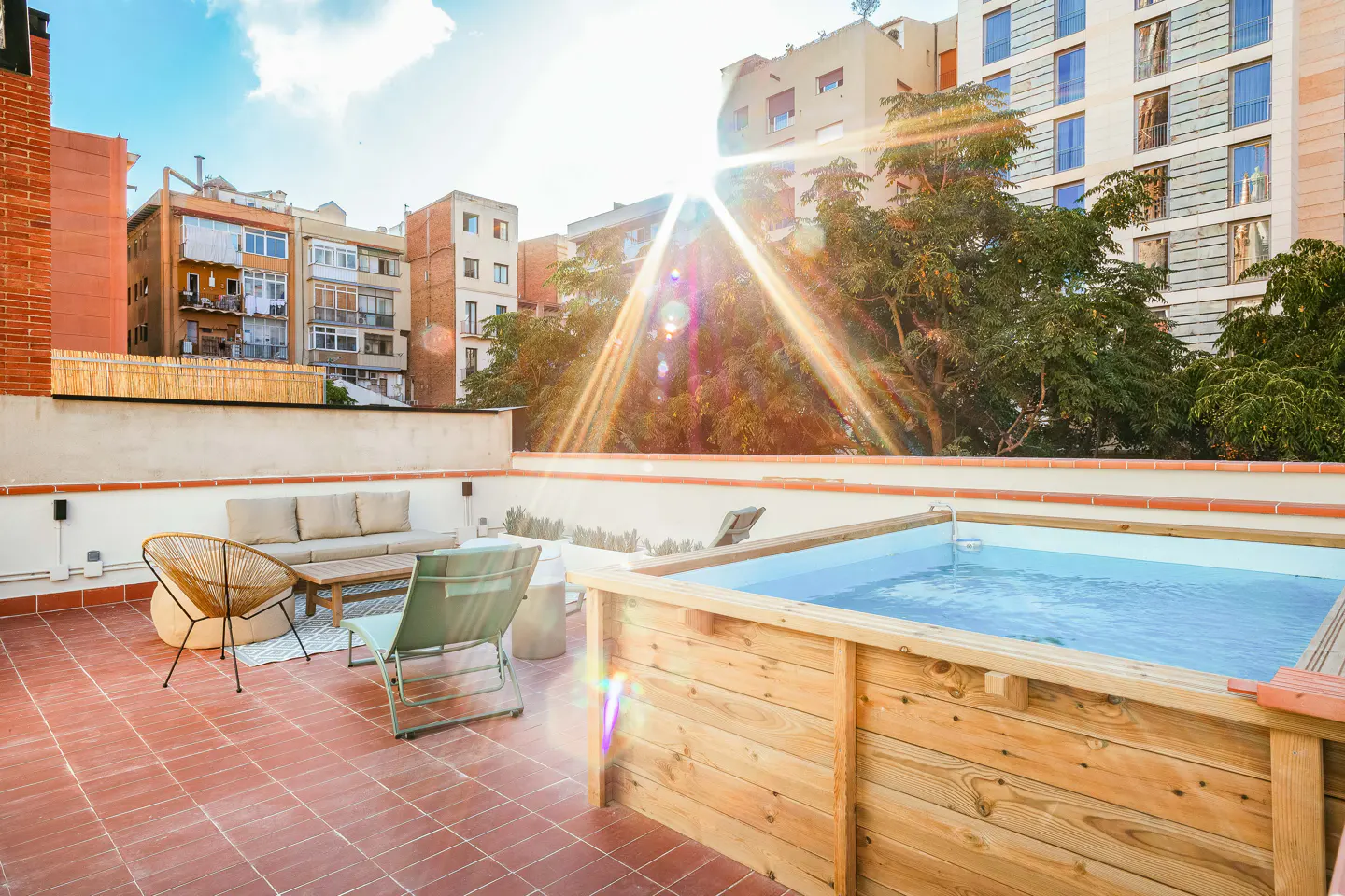 Rooftop patio with a small wooden pool, lounge seating, and city views under a sunny sky.