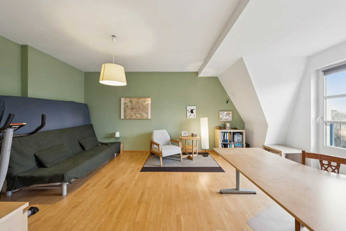 Bright living space with green walls, wood floors, and white ceiling. Features a futon, chair, desk, and exercise bike.