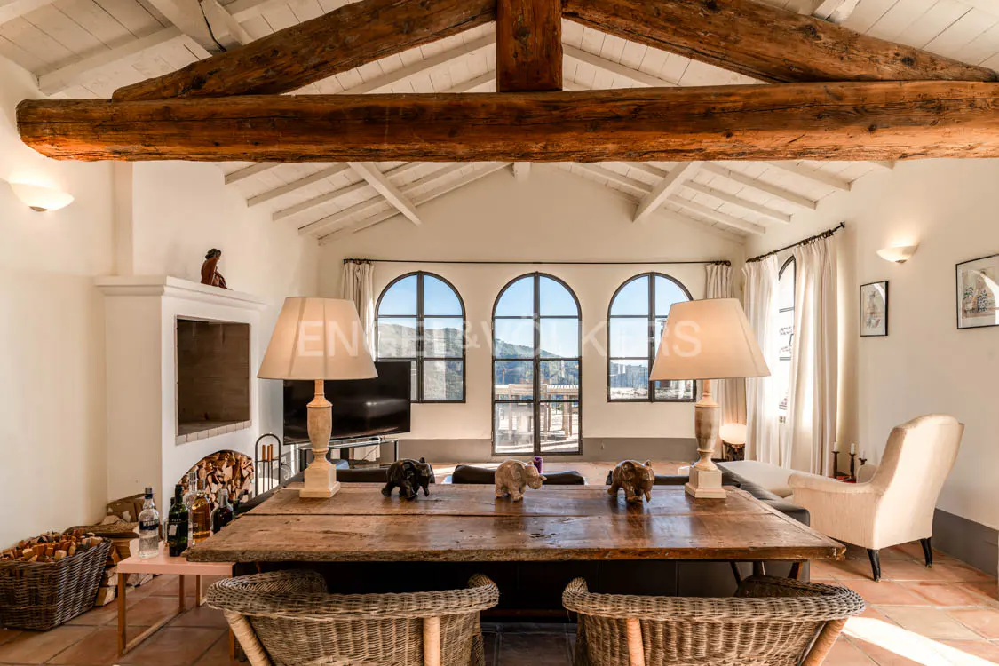Bright living room with exposed beams, fireplace, and arched windows. A rustic wooden table sits in the center with wicker chairs.