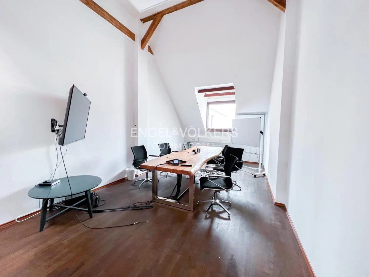 Attic office space with a live edge conference table, black chairs, and a wall-mounted TV. White walls and dark wood floors.