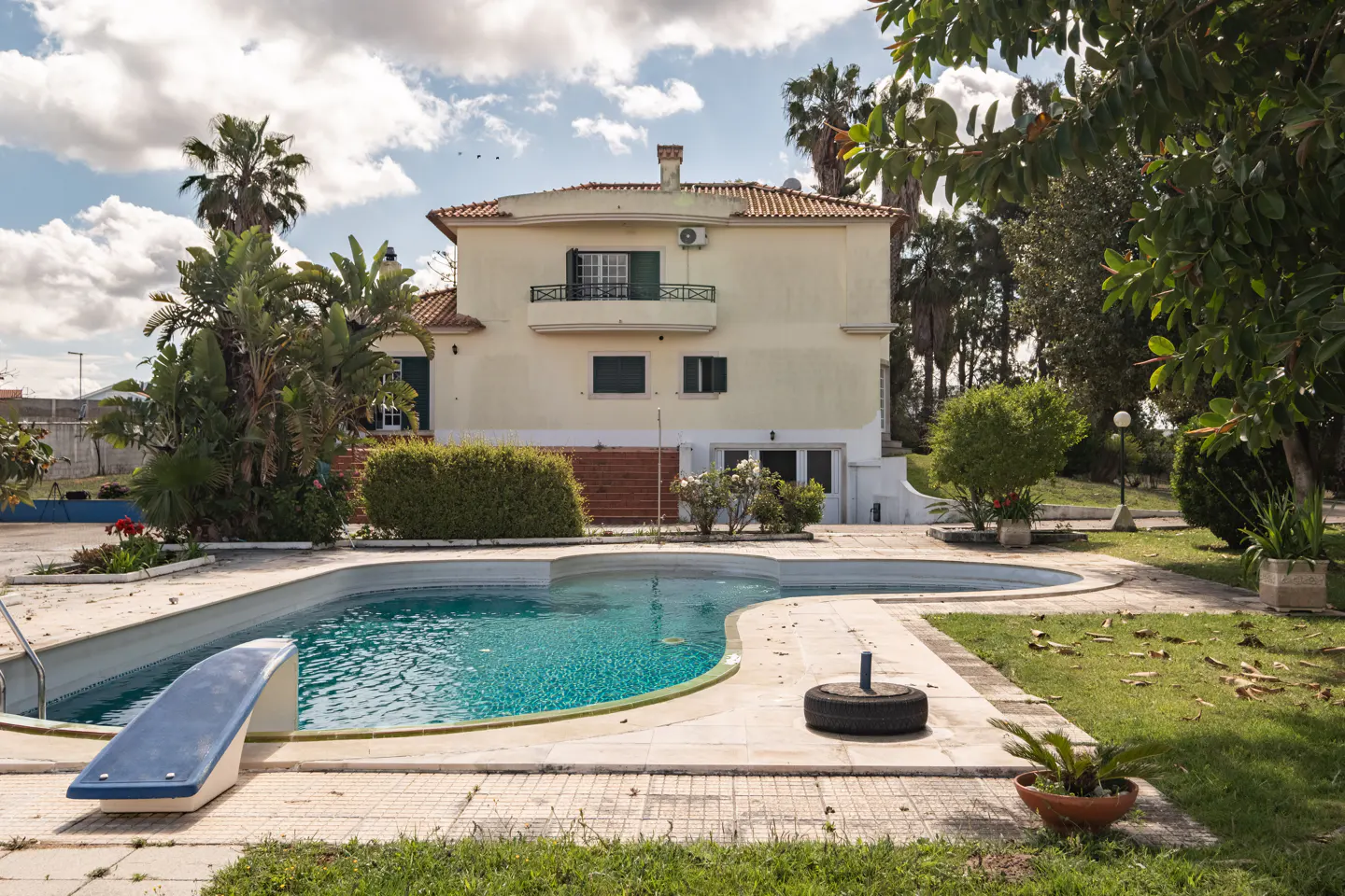 Two-story beige house with a red tile roof, a blue swimming pool, and palm trees under a cloudy sky.