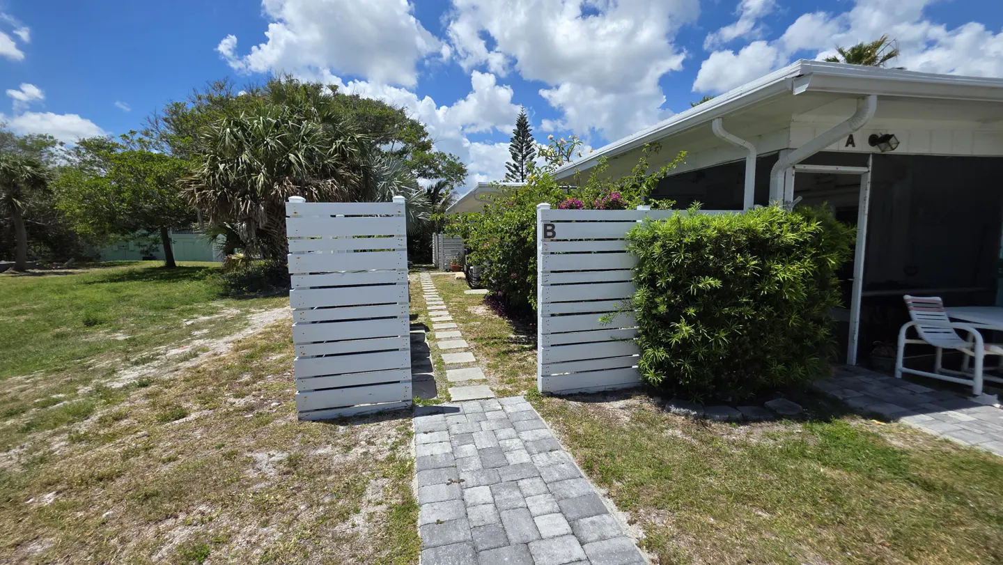 Exterior view of a white beach house with a stone walkway and white wooden fence. Unit B is visible. Blue sky with clouds.