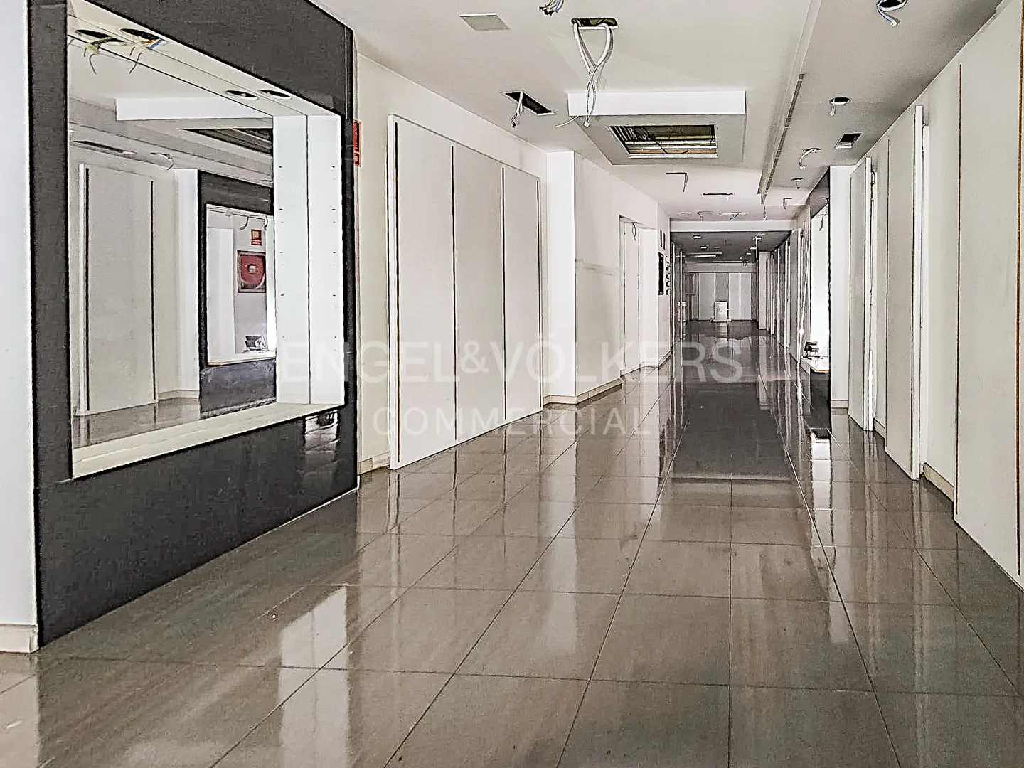 A long, empty commercial hallway with white walls and gray tiled floors. A black framed mirror is on the left.
