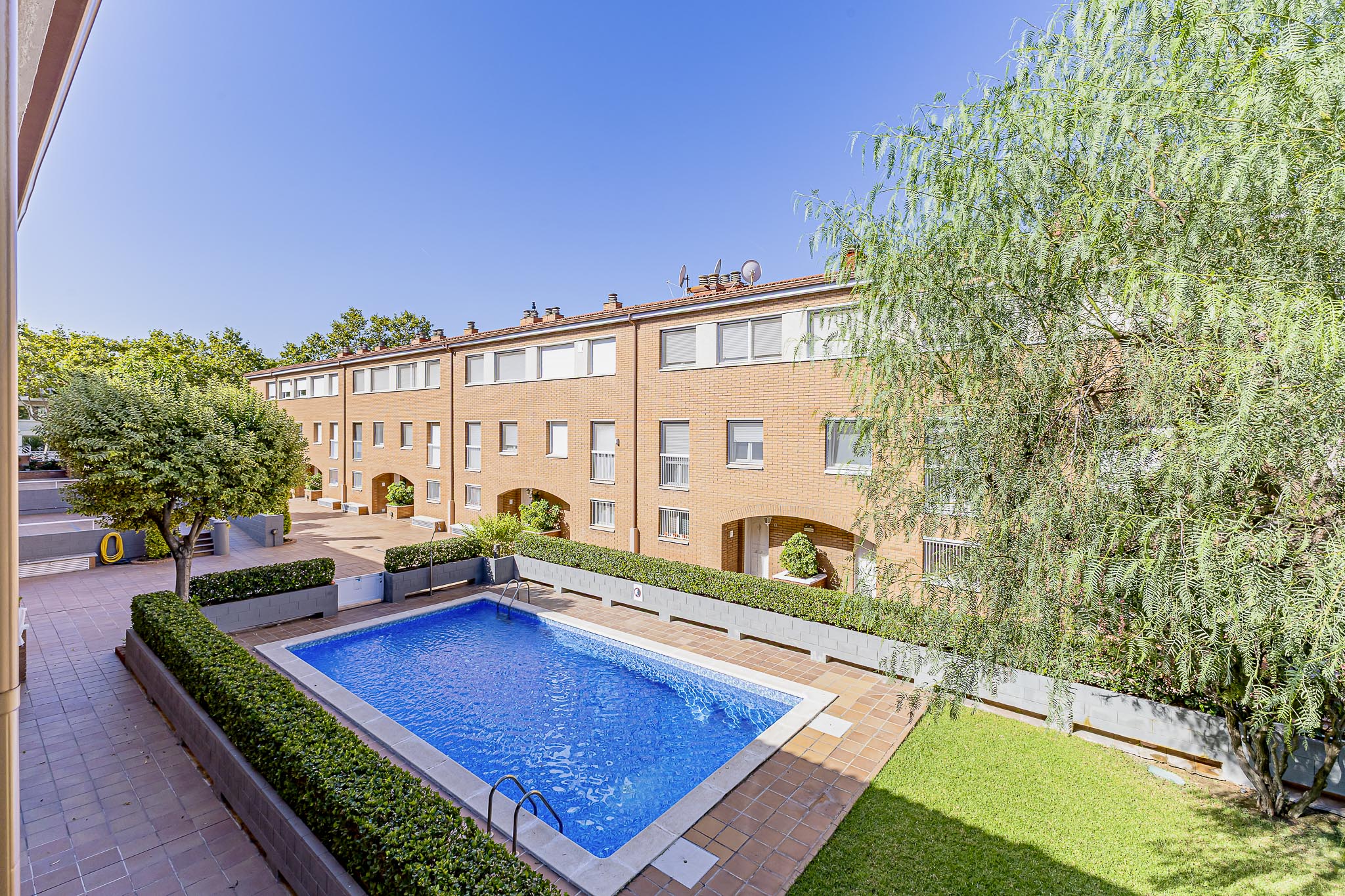 A rectangular swimming pool with blue water sits in a courtyard surrounded by brick townhouses and manicured hedges. [AI-generated text]