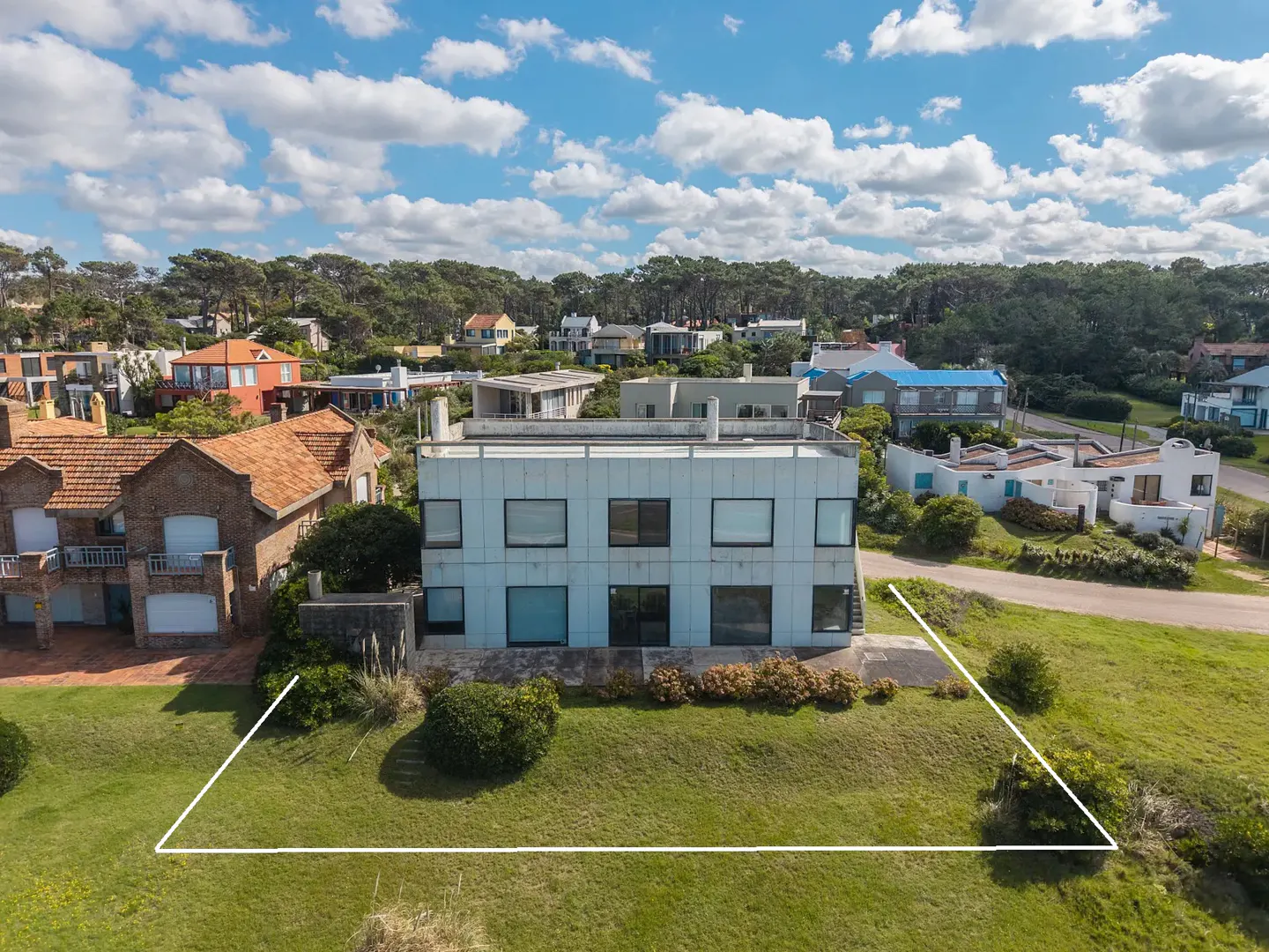 Aerial view of a modern, two-story white house with a flat roof, surrounded by green grass and other houses. Blue sky with clouds.