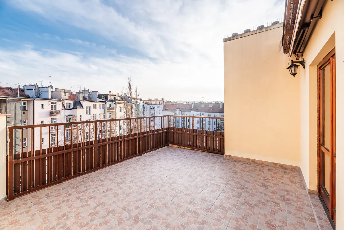A rooftop balcony with brown wood railing and tiled floor overlooks a city with white buildings under a blue sky.