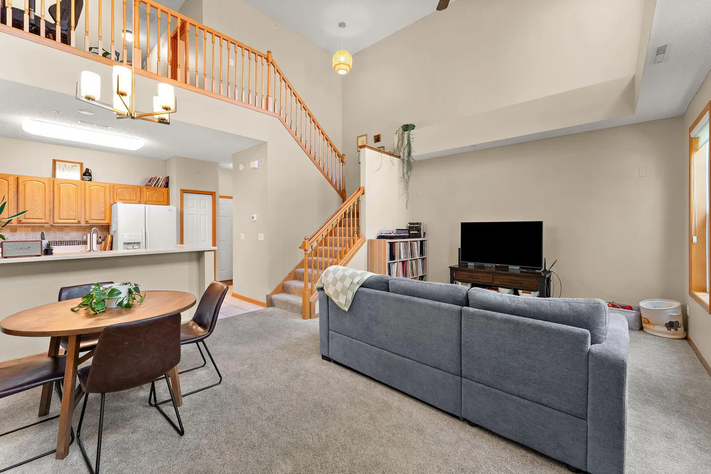 Open-concept living room with a gray sofa, wood dining table, and staircase leading to a loft. The kitchen has wood cabinets and white appliances.