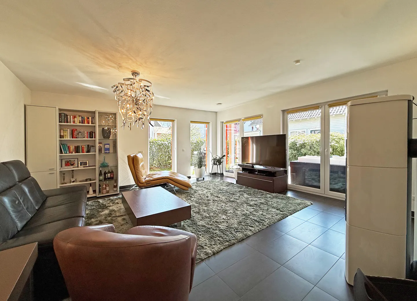Bright living room with a black leather sofa, brown leather chair, and a chaise lounge. A large TV and bookshelf are also visible.