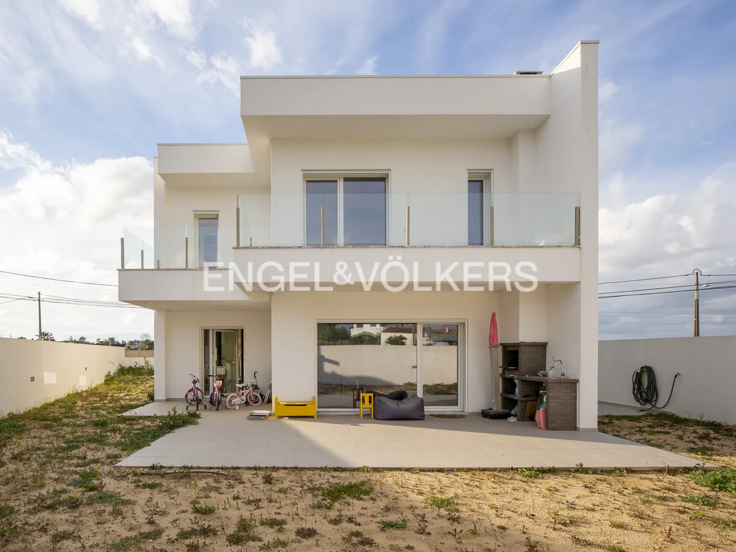 Modern two-story white house with a patio, bikes, and outdoor kitchen. Engel & Völkers logo is visible.