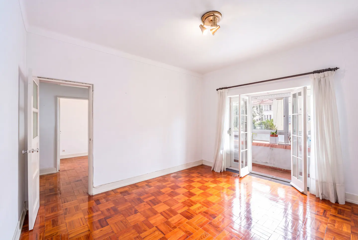 Bright, empty room with parquet floor, white walls, and open French doors to a balcony with a plant.