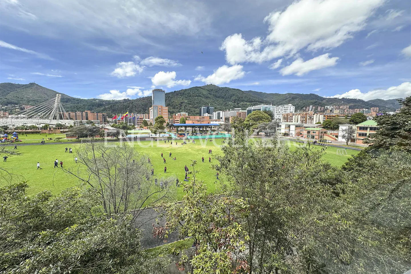 A wide view of a green park with people walking, framed by trees, with a city skyline and mountains in the background.
