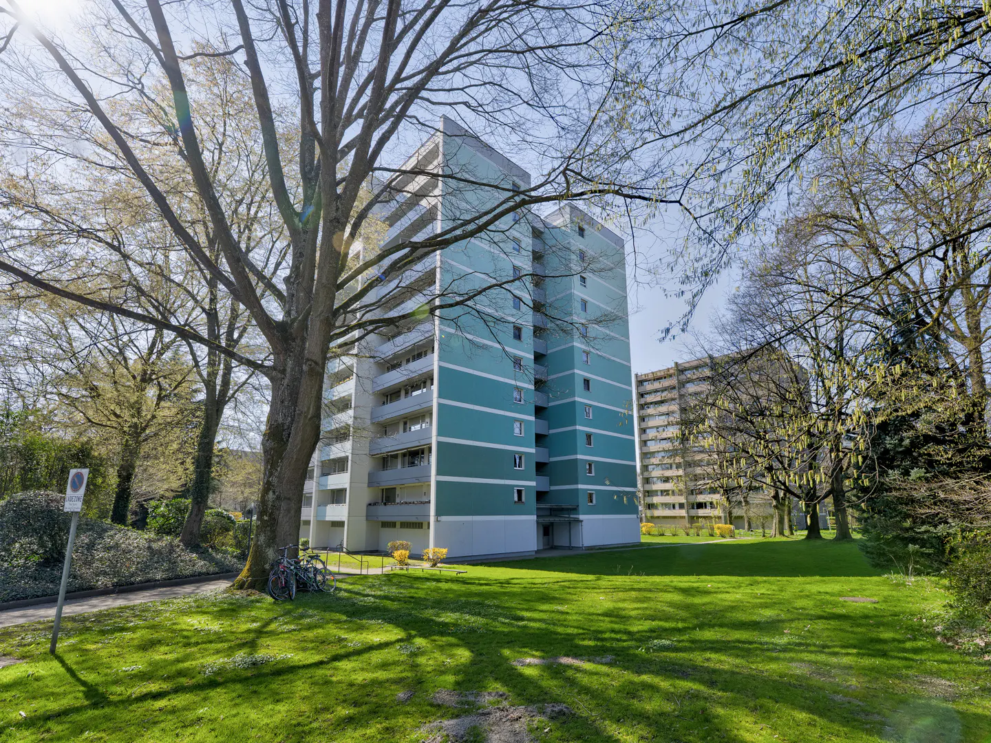 Exterior view of a tall, blue and white apartment building with balconies, surrounded by green grass and trees.