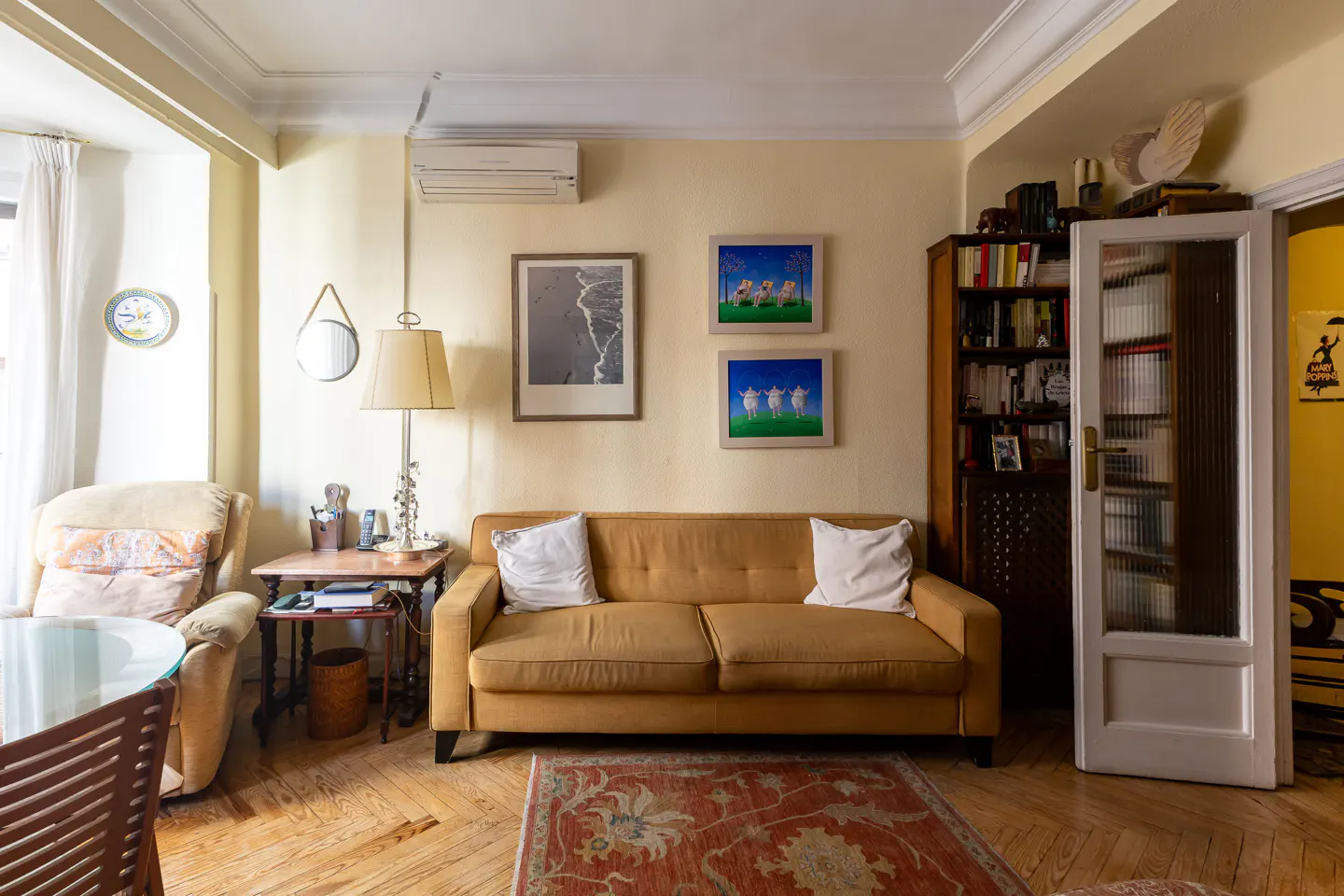 Living room with a yellow sofa, white pillows, and a red patterned rug on a wood floor. Art hangs above the sofa. A bookcase and glass door are to the right.