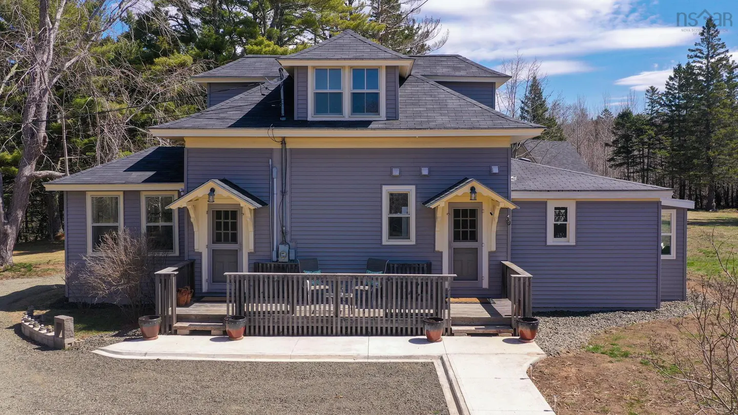 Two-story gray house with a black roof, yellow trim, and a wooden deck. Two doors with small awnings face the front. Trees in the background.