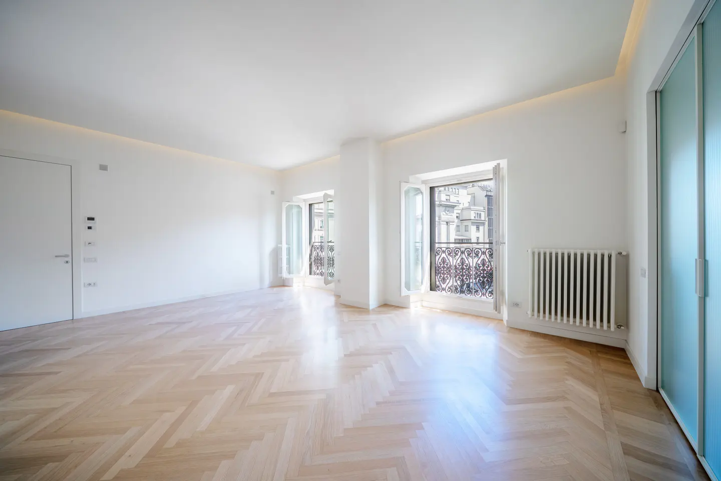 Bright, empty room with herringbone wood floors, white walls, and two open windows with wrought iron balconies. A radiator sits below one window.