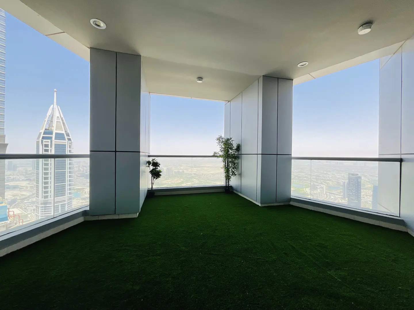 A high-rise balcony with artificial grass, potted plants, and a city view through a glass railing.