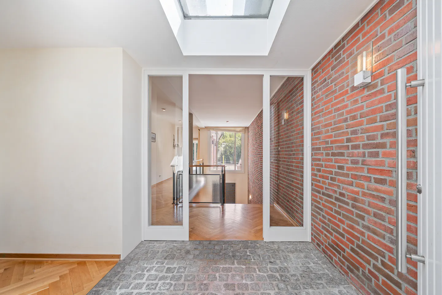Modern entryway with a cobblestone floor, red brick wall, and glass doors leading to a bright hallway with a skylight.