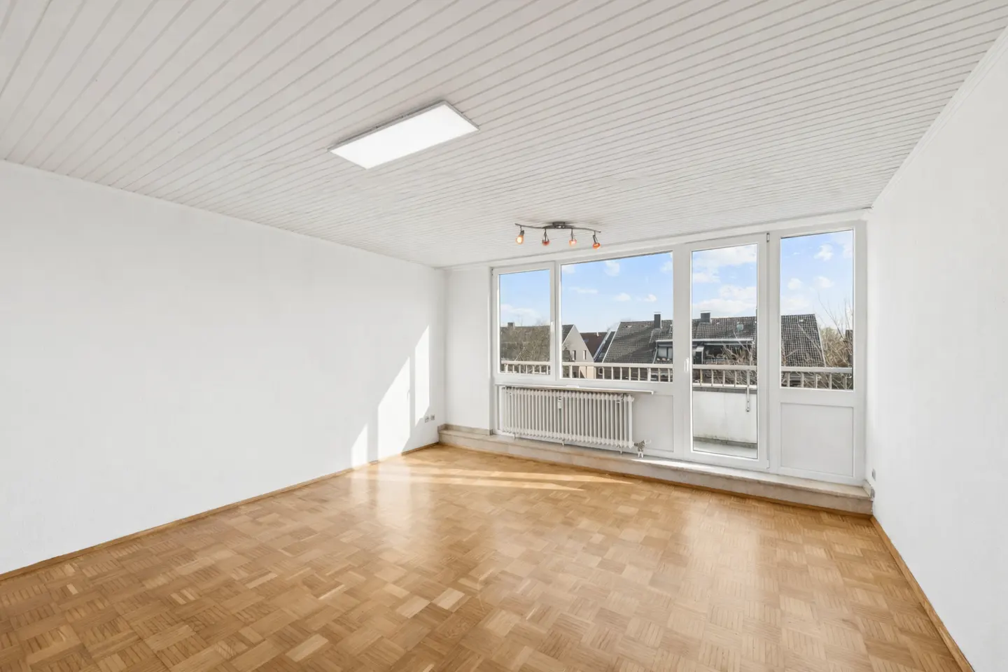 Bright, empty room with parquet floor, white walls, and a large window with a balcony overlooking rooftops. A radiator sits beneath the window.