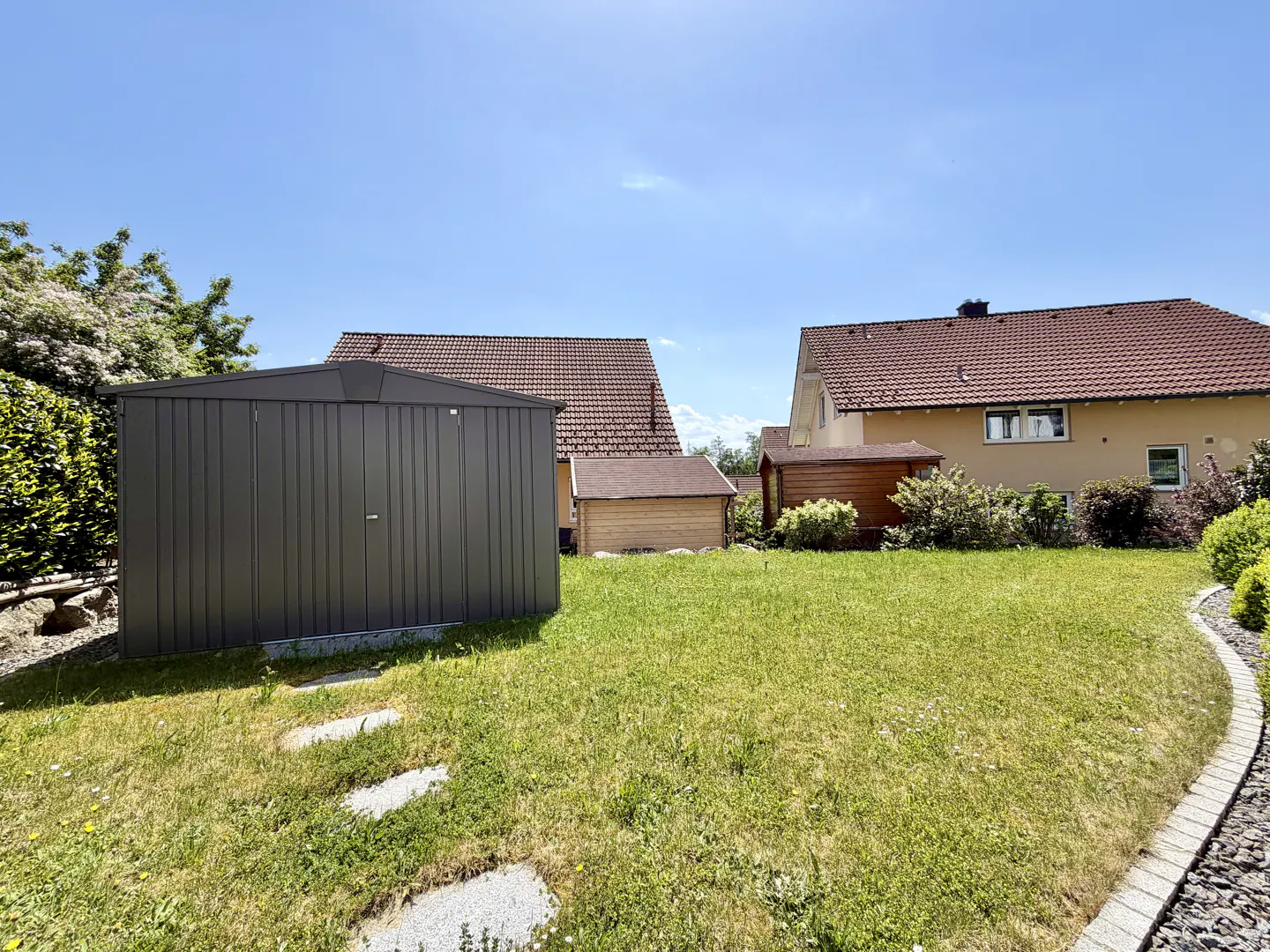 A dark grey metal storage shed sits on a grassy backyard lawn with houses and trees in the background under a blue sky.