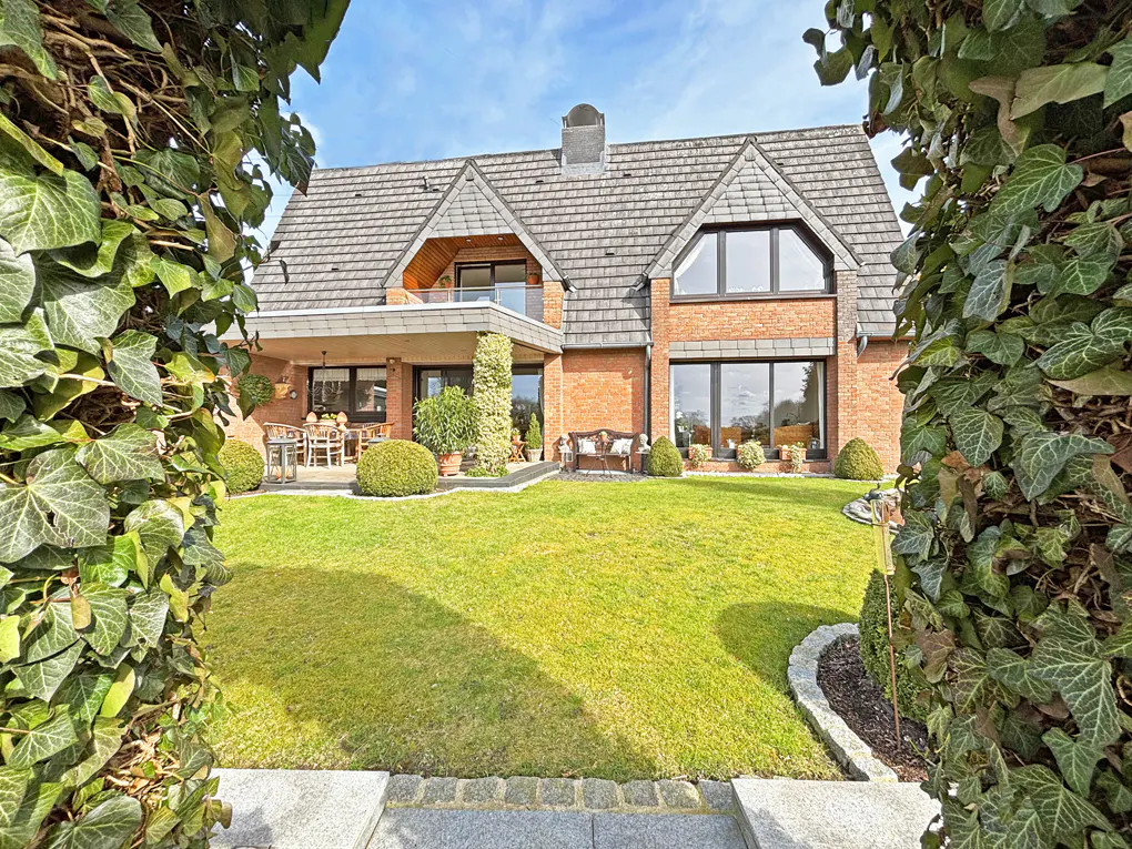 View of a red brick house with a gray roof and green lawn, framed by ivy. Patio furniture and a bench are visible.