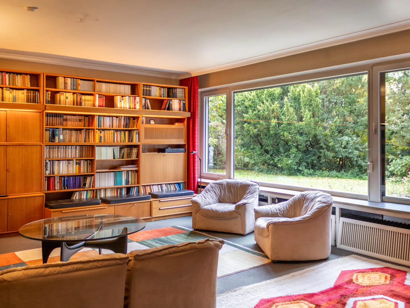 A living room with a large bookshelf, two armchairs, and a large window with a view of trees.
