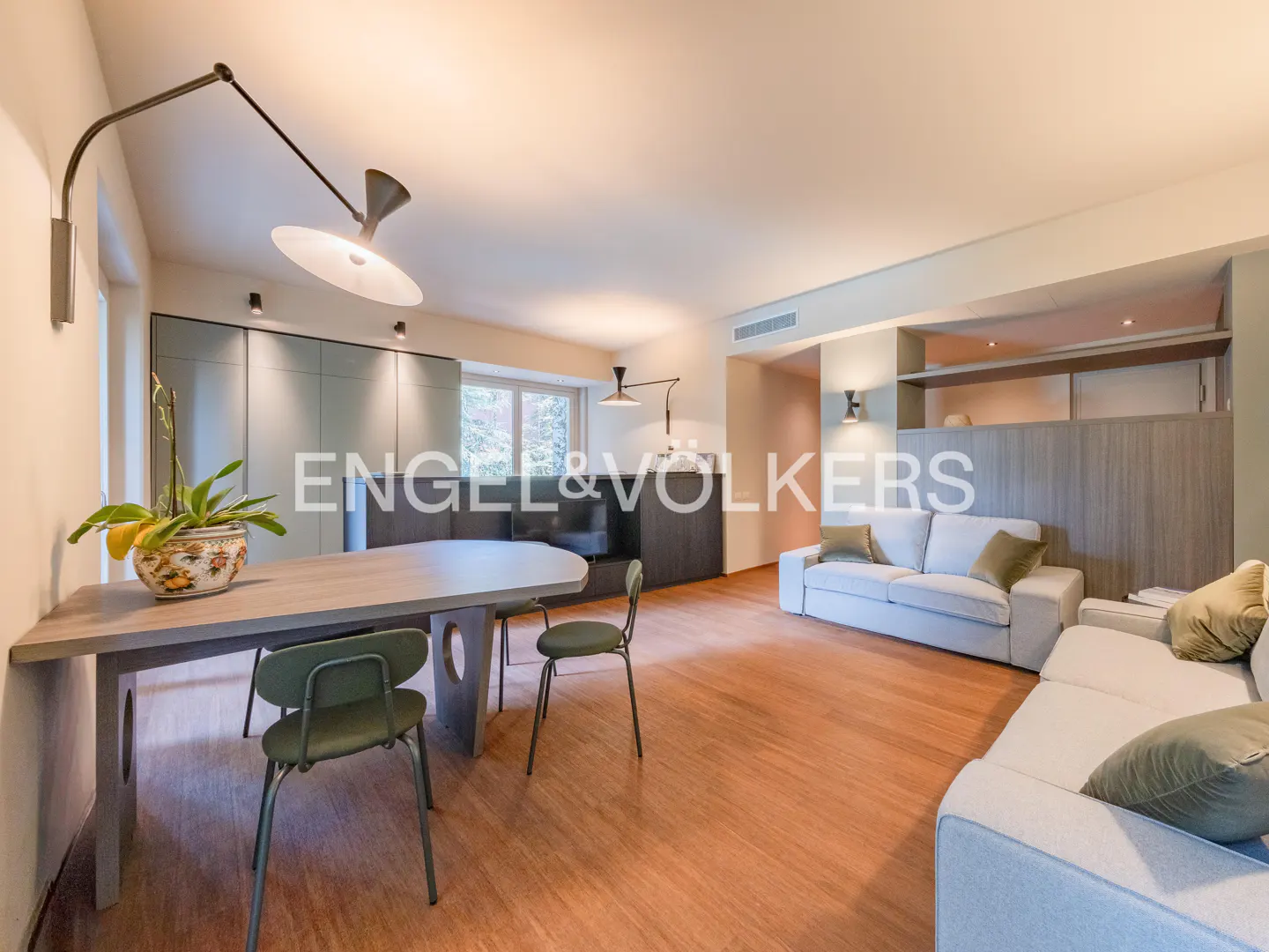 Open-concept living space with wood floors, a dining table with green chairs, and a white sofa. Modern lighting fixtures illuminate the room.