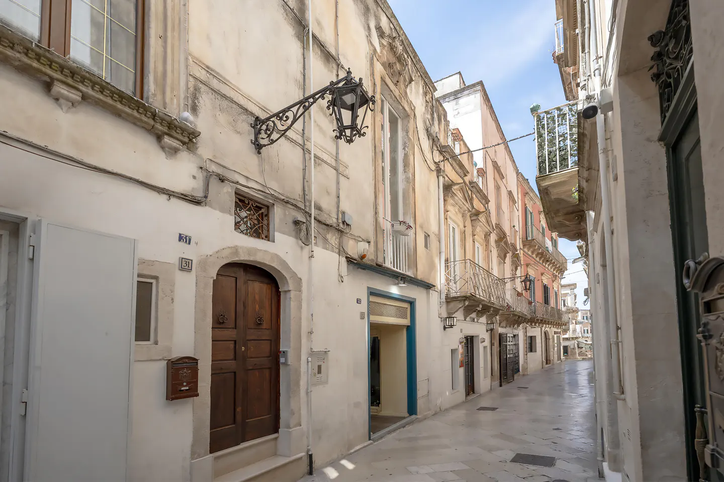 Narrow street view of old buildings with balconies and a brown wooden door on the left.