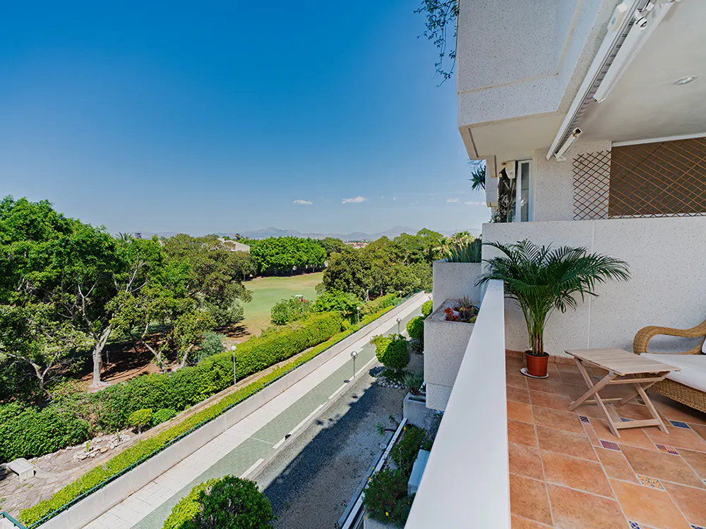 Balcony view of a golf course. A palm tree sits on the tiled balcony with a table and chair. Green trees and blue sky in the background.