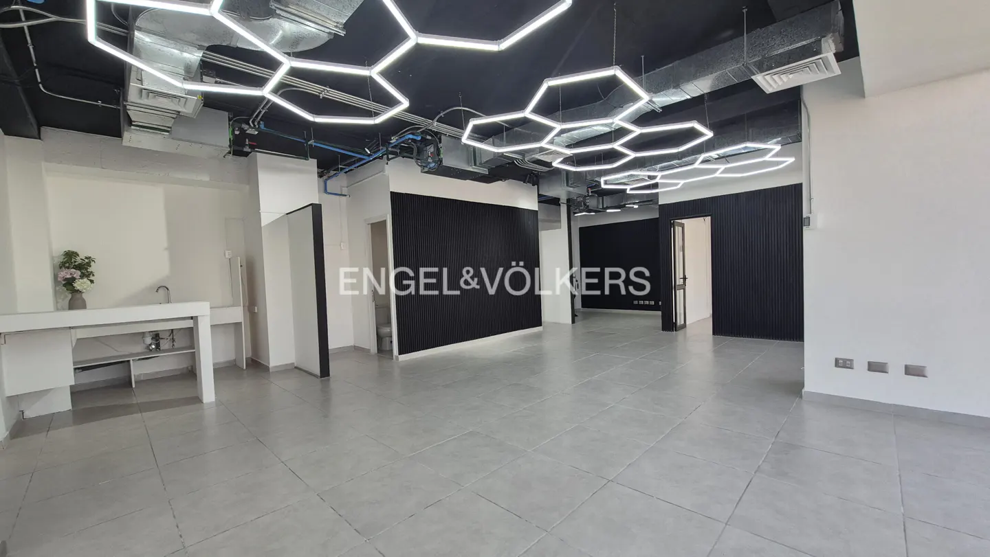 Empty office space with gray tile floors, white walls, and black accents. Hexagonal lights hang from the black ceiling. Engel & Völkers logo is visible.