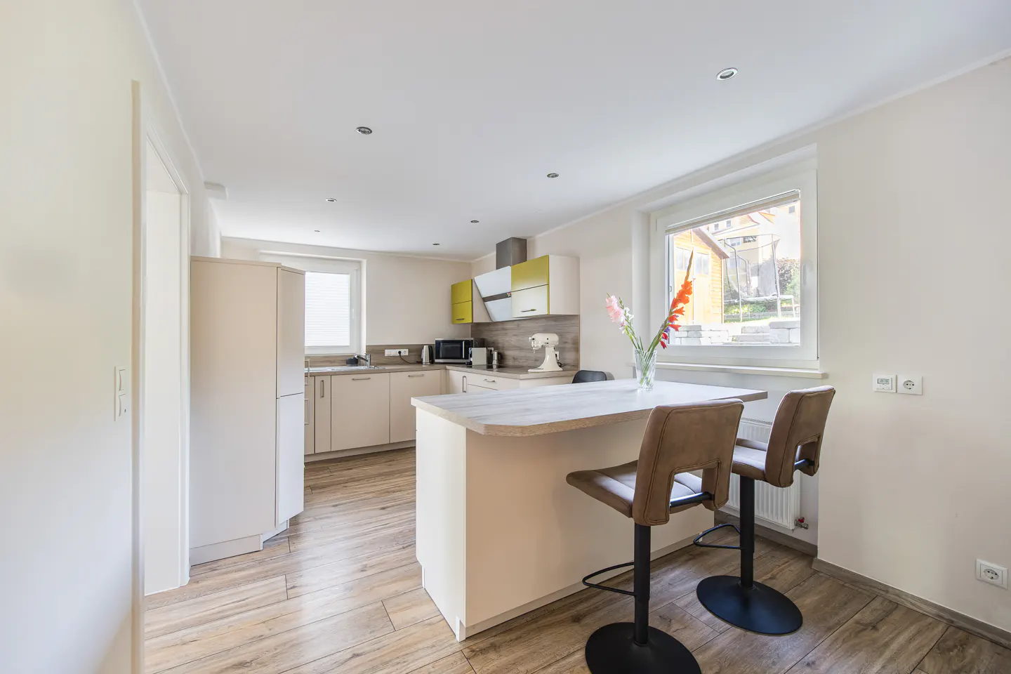 Bright kitchen with wood floors, white cabinets, and a breakfast bar with two brown leather stools. Flowers in a vase sit on the counter by a window.