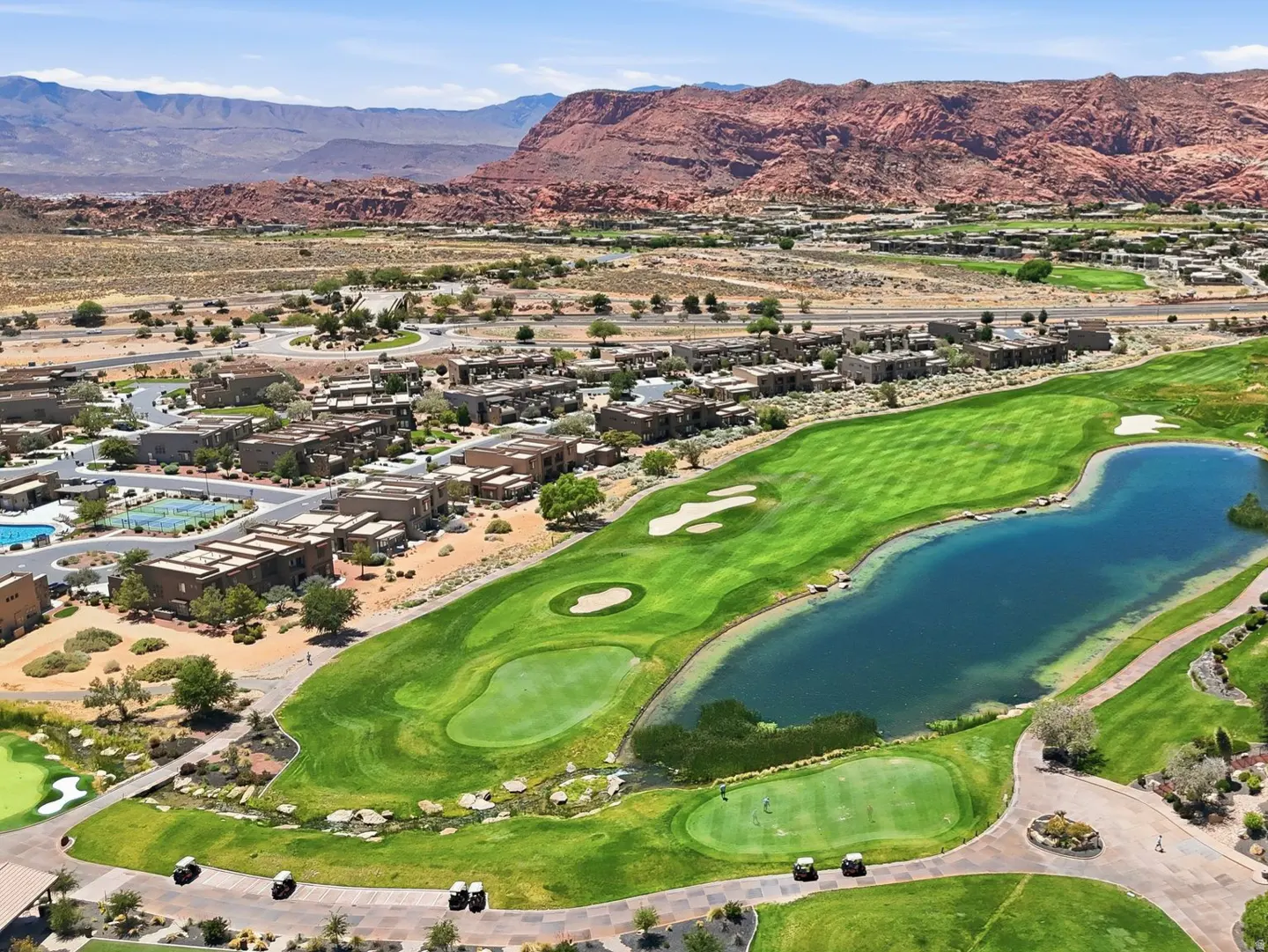 Aerial view of a lush green golf course with a large blue pond, surrounded by desert homes and red rock mountains.