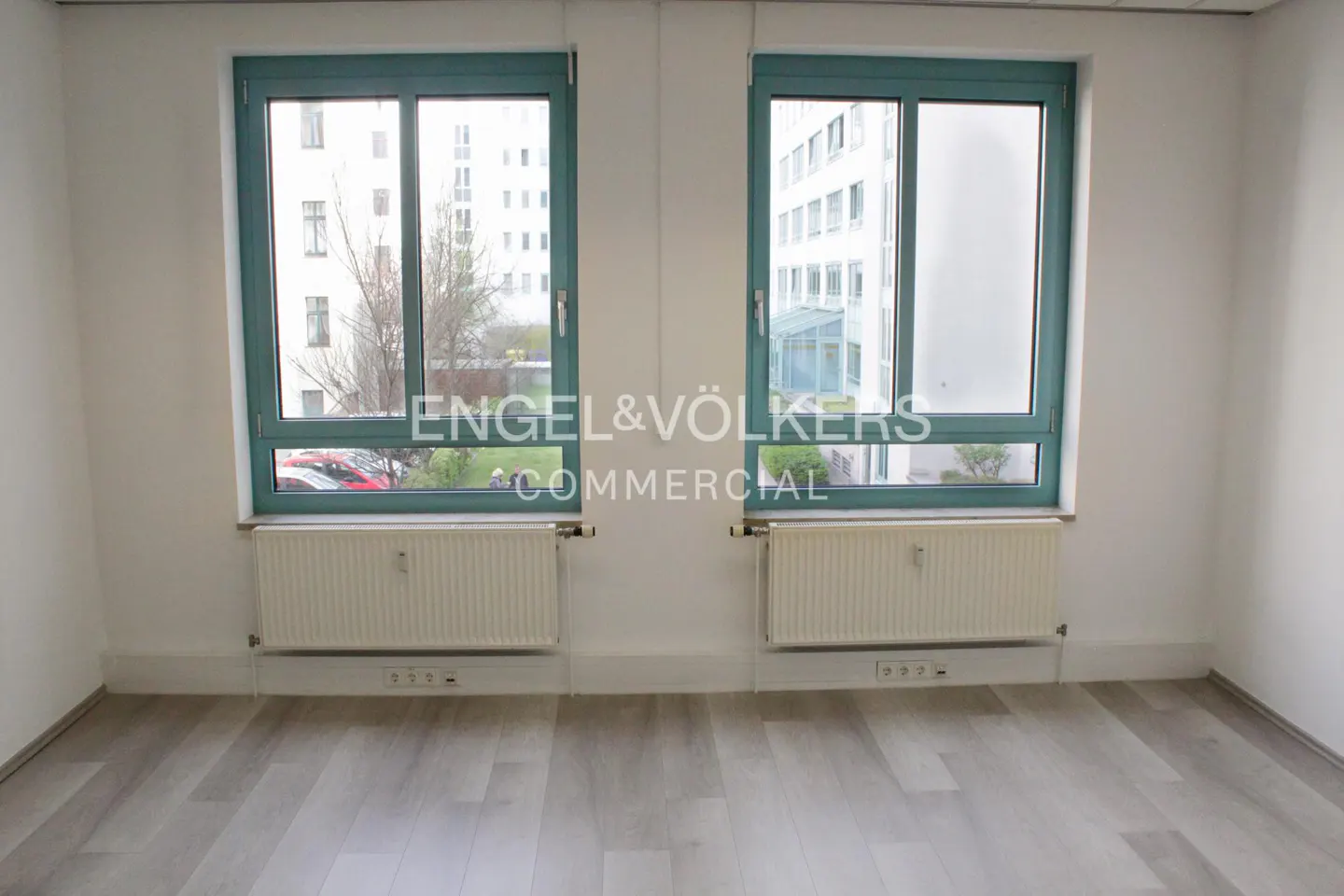 Empty room with gray wood floors, white walls, and two teal-framed windows overlooking a courtyard. Radiators sit beneath each window.