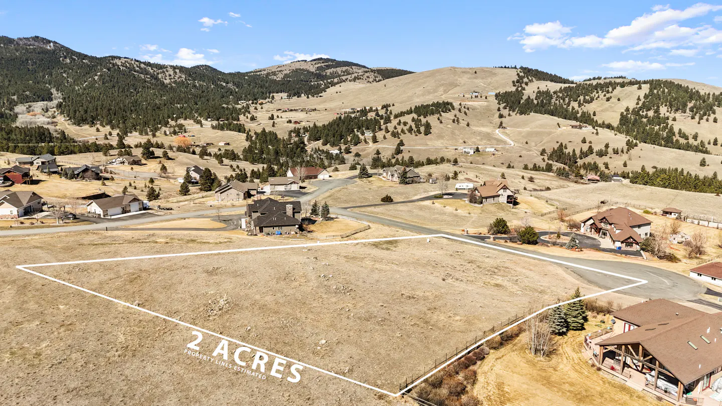 Aerial view of a 2-acre lot outlined in white, nestled in a hilly landscape with scattered houses and trees under a blue sky.