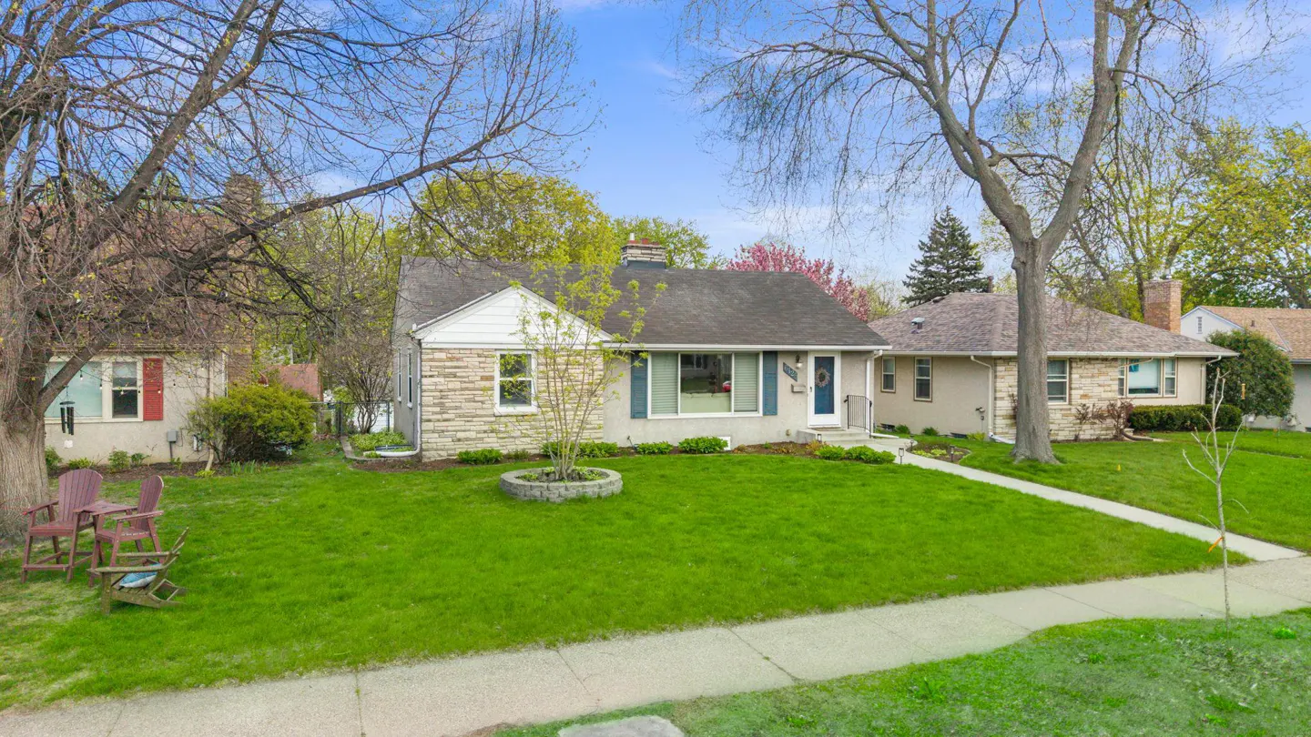 A single-story house with a stone and stucco exterior, a dark roof, and a green lawn on a sunny day.