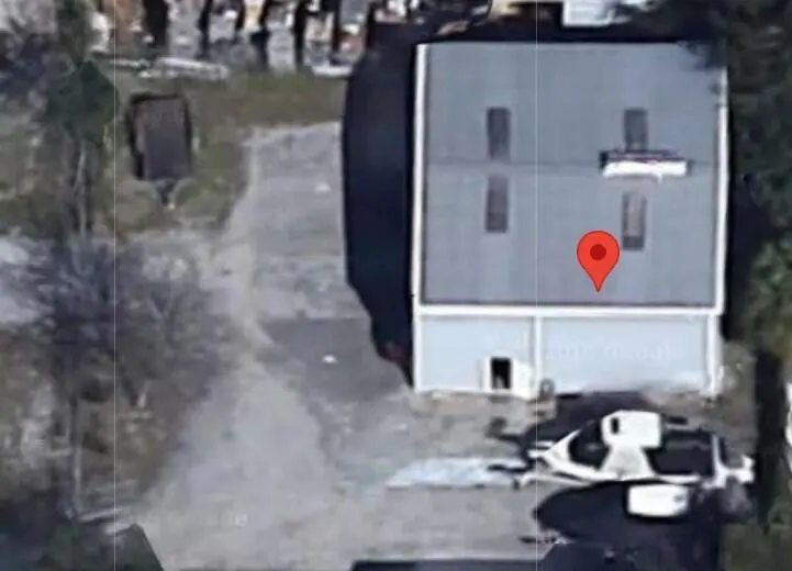 Aerial view of a gray commercial building with a red location pin on the roof, surrounded by a parking lot and trees.