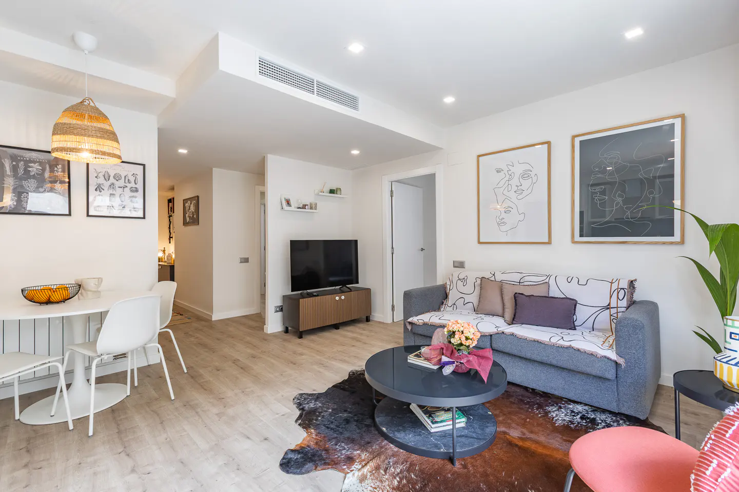 Bright living room with a gray sofa, round coffee table, and dining area with white chairs. Artwork adorns the white walls.