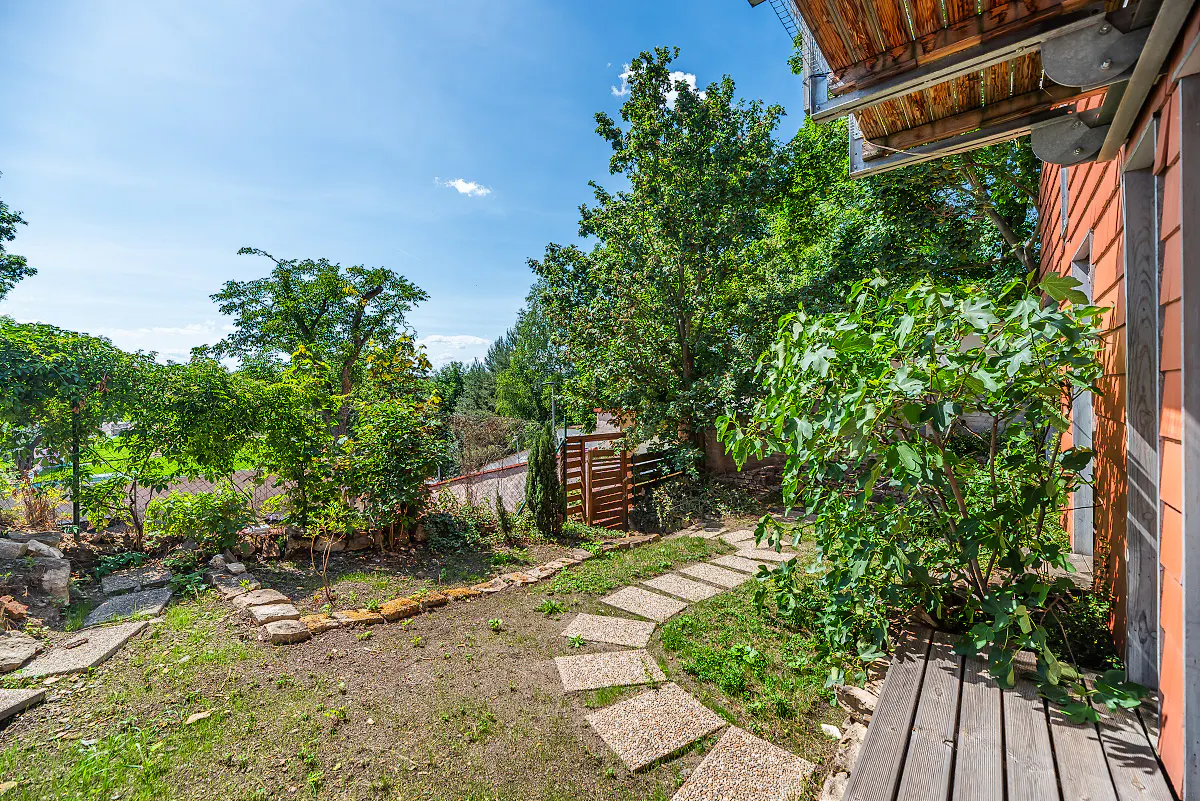 A stone path winds through a sunny backyard with lush green trees, a wooden deck, and a terracotta-tiled house exterior.