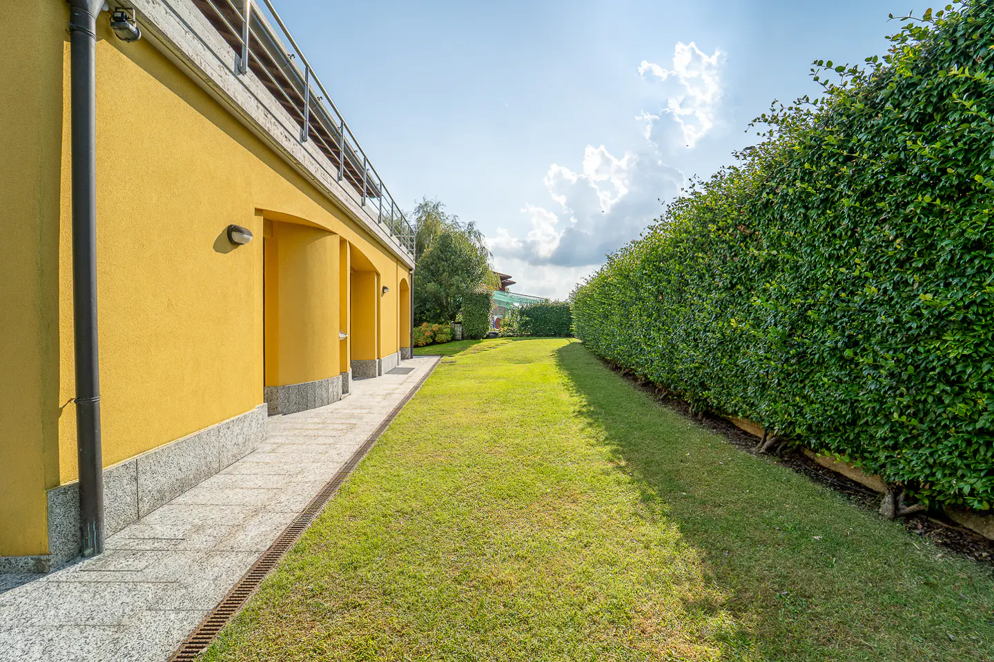A yellow building with arched doorways borders a green lawn, next to a tall, green hedge under a blue sky.