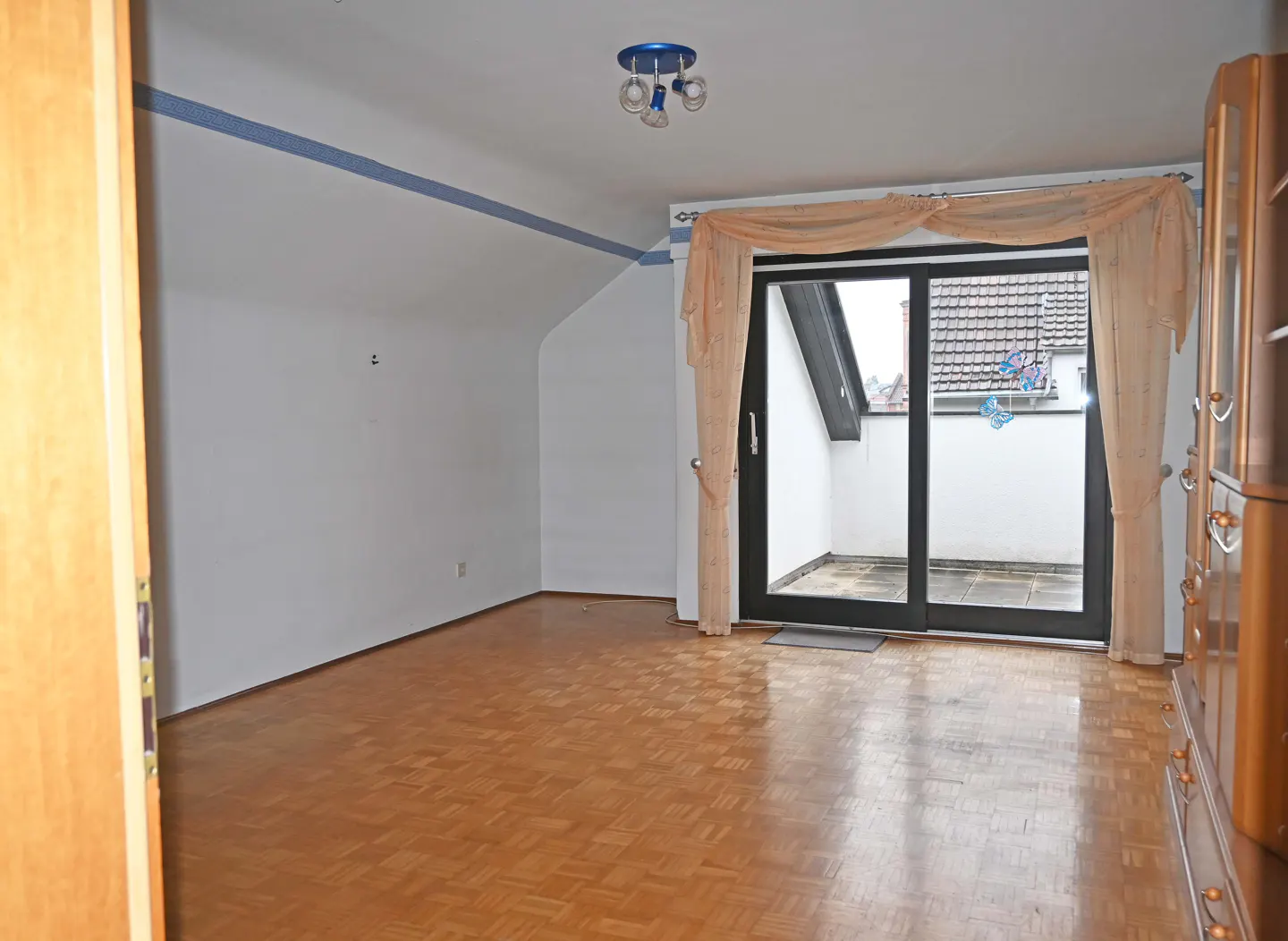 A bright room with wood floors, white walls, and a blue stripe near the ceiling. A glass door leads to a balcony with a view of rooftops.