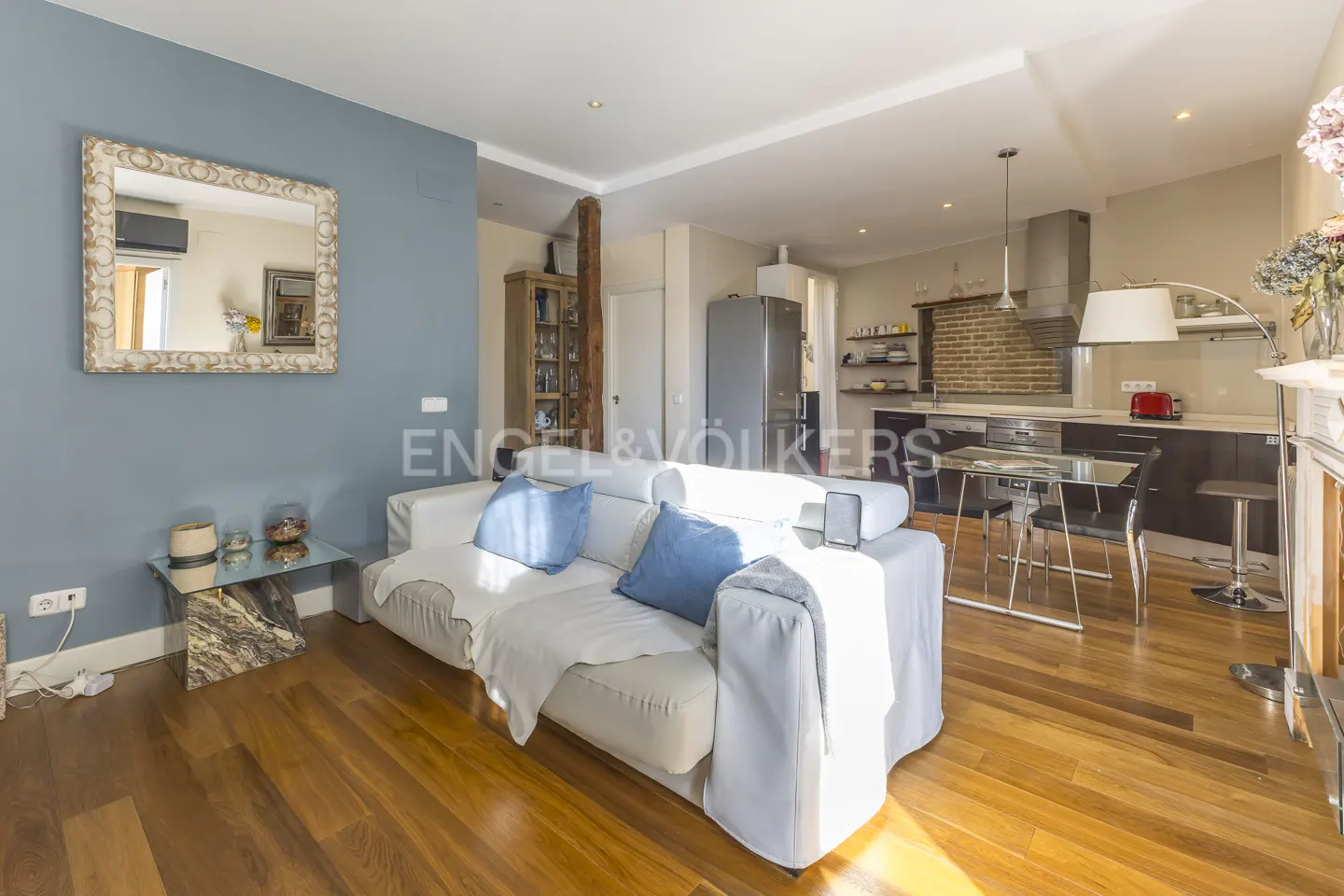 Open-concept living space with hardwood floors, a white sofa with blue pillows, and a modern kitchen in the background.